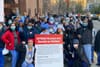 A group of more than two dozen nurses in scrubs stand together in front of a hospital, with some clutching a sign that reads,