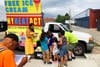 A bunch of kids in front of a white truck wait for popsicles to be handed out from a woman in a yellow dress, while a man in